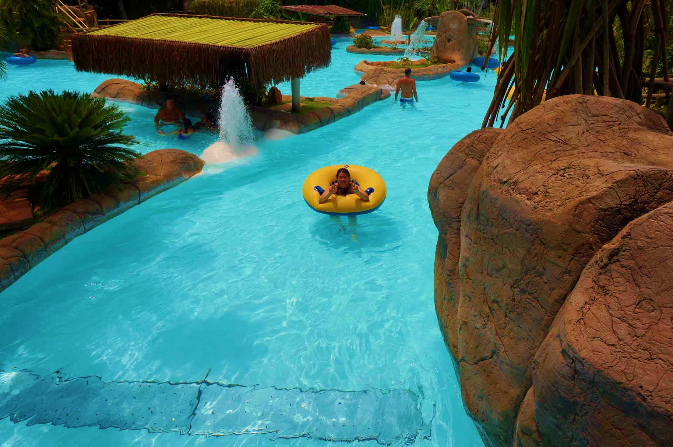Family enjoying tropical water park with turquoise pool, yellow inflatable float, rock formations, and thatched-roof cabana i